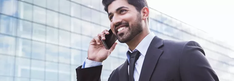 Person in a suit smiling while talking on a phone in front of a glass building.
